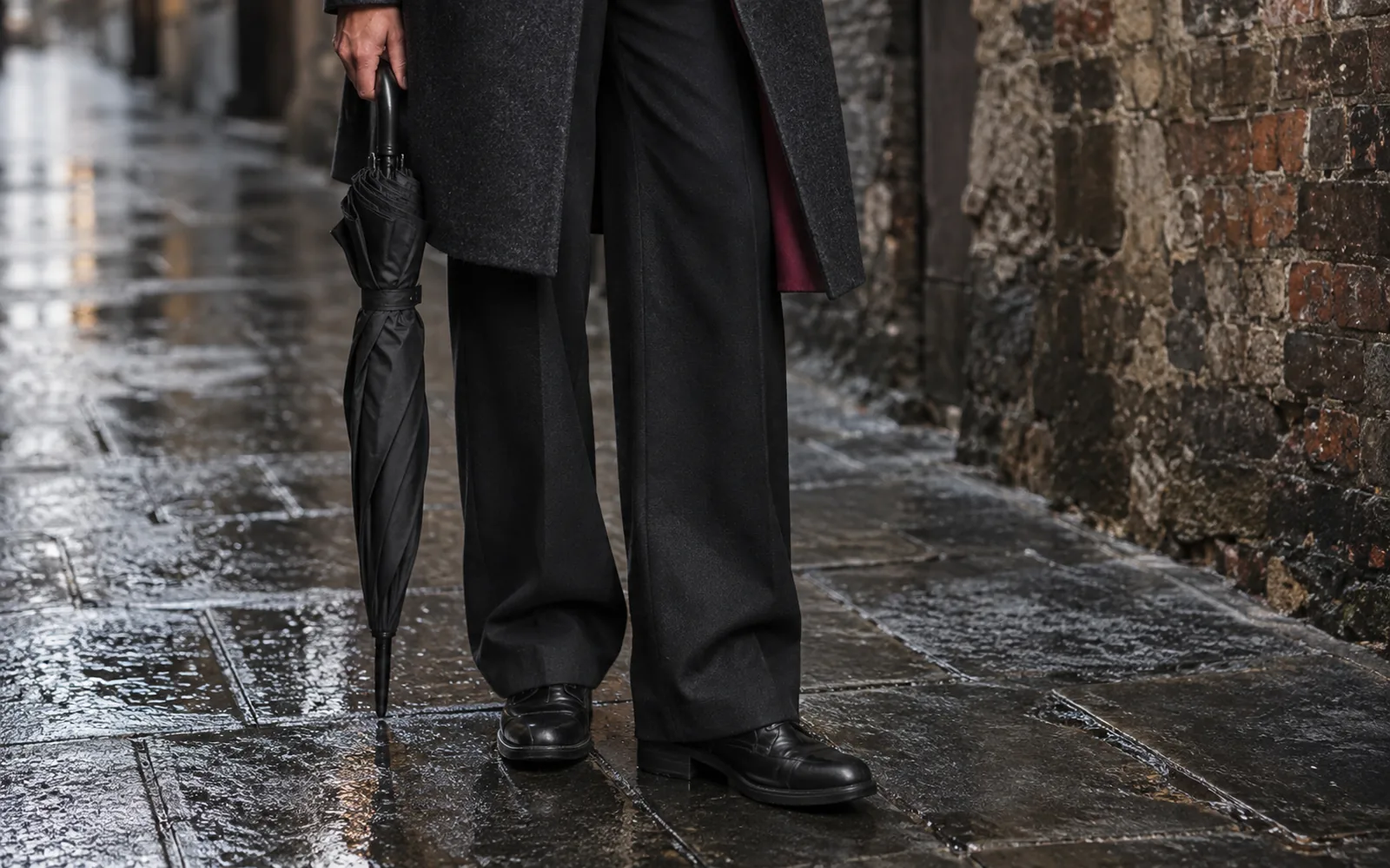 Illustrative close crop of wide black trousers, a long dark coat hem and leather boots on a damp bluestone laneway, with a folded umbrella
