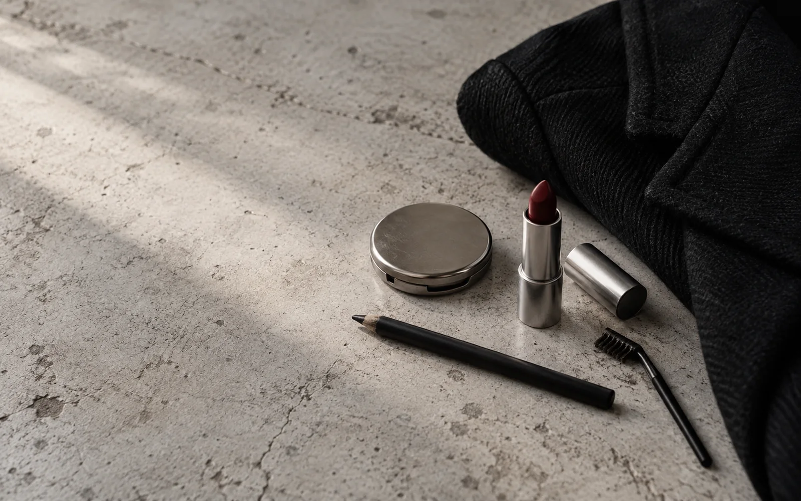 Illustrative still life of unbranded makeup on concrete beside a folded black jacket: an eyeliner pencil, a lipstick, a small compact and a brow brush