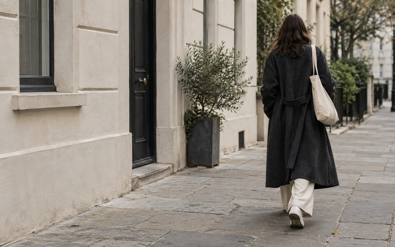 Illustrative street scene: an anonymous person seen from behind in a long coat and wide trousers walking along a city pavement